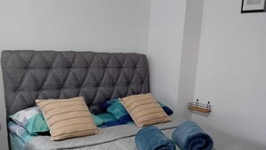 Un lit bien fait, recouvert d’une courtepointe matelassée grise, avec deux serviettes bleues enroulées et plusieurs oreillers de différentes couleurs et motifs.