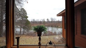 A window with a view of a lake, a wooden structure, and a potted plant on a stand. - Cozy 3-bedroom cabin in charming Crosslake with WiFi and AC (Crosslake)