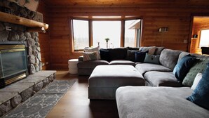 A cozy cabin interior with a stone fireplace, a sectional sofa, and large windows offering a view of the outdoors. - Cozy 3-bedroom cabin in charming Crosslake with WiFi and AC (Crosslake)