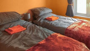 Une chambre avec deux lits, chacun étant équipé de draps gris et de serviettes orange. Une lampe se trouve sur la table de chevet à côté de l’un des lits.