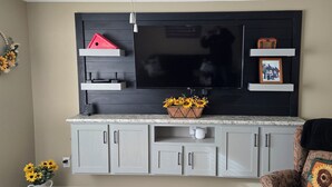 Een woonkamer met een grote flatscreen televisie aan een donkere muur, grijze kastjes eronder en een klein tafeltje met een mand vol bloemen.