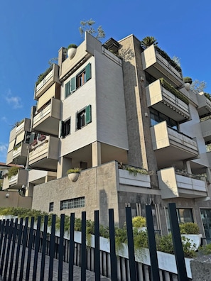 A multi-story building with balconies, a stone facade, and a black fence in the foreground.