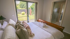 Ein Schlafzimmer mit Bett, Kissen, einer Nachttischlampe, einem Badezimmer mit Glastür und Blick auf einen Garten.