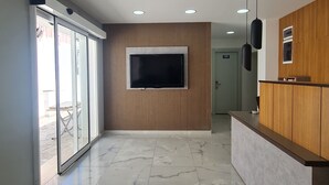 Un intérieur moderne avec un grand téléviseur fixé sur un mur recouvert de panneaux en bois, un comptoir d’accueil doté de meubles en bois et une porte en verre donnant sur l’extérieur.
