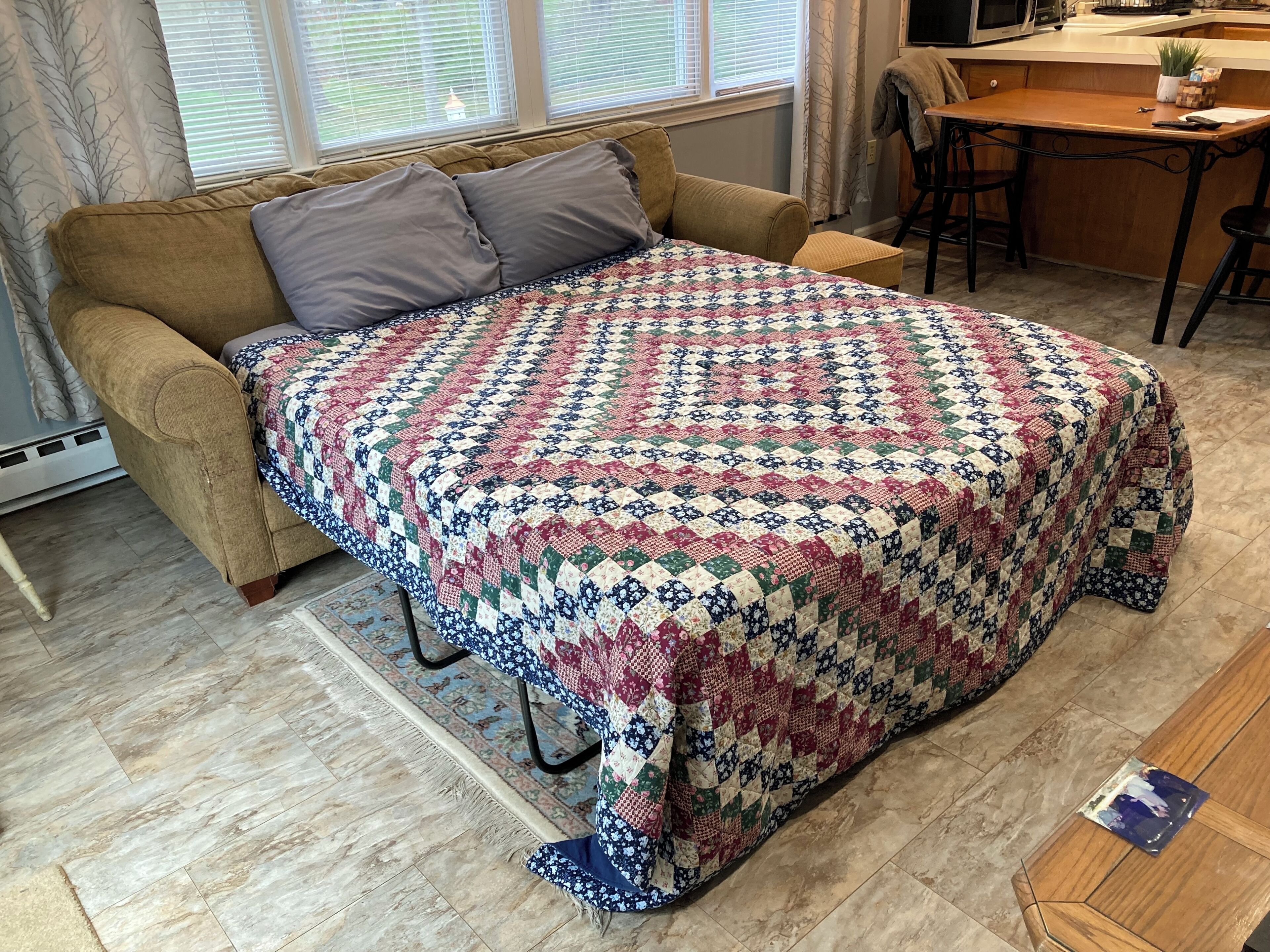 A cozy room with a brown sofa, a patterned quilt, a wooden coffee table, and a window with blinds.