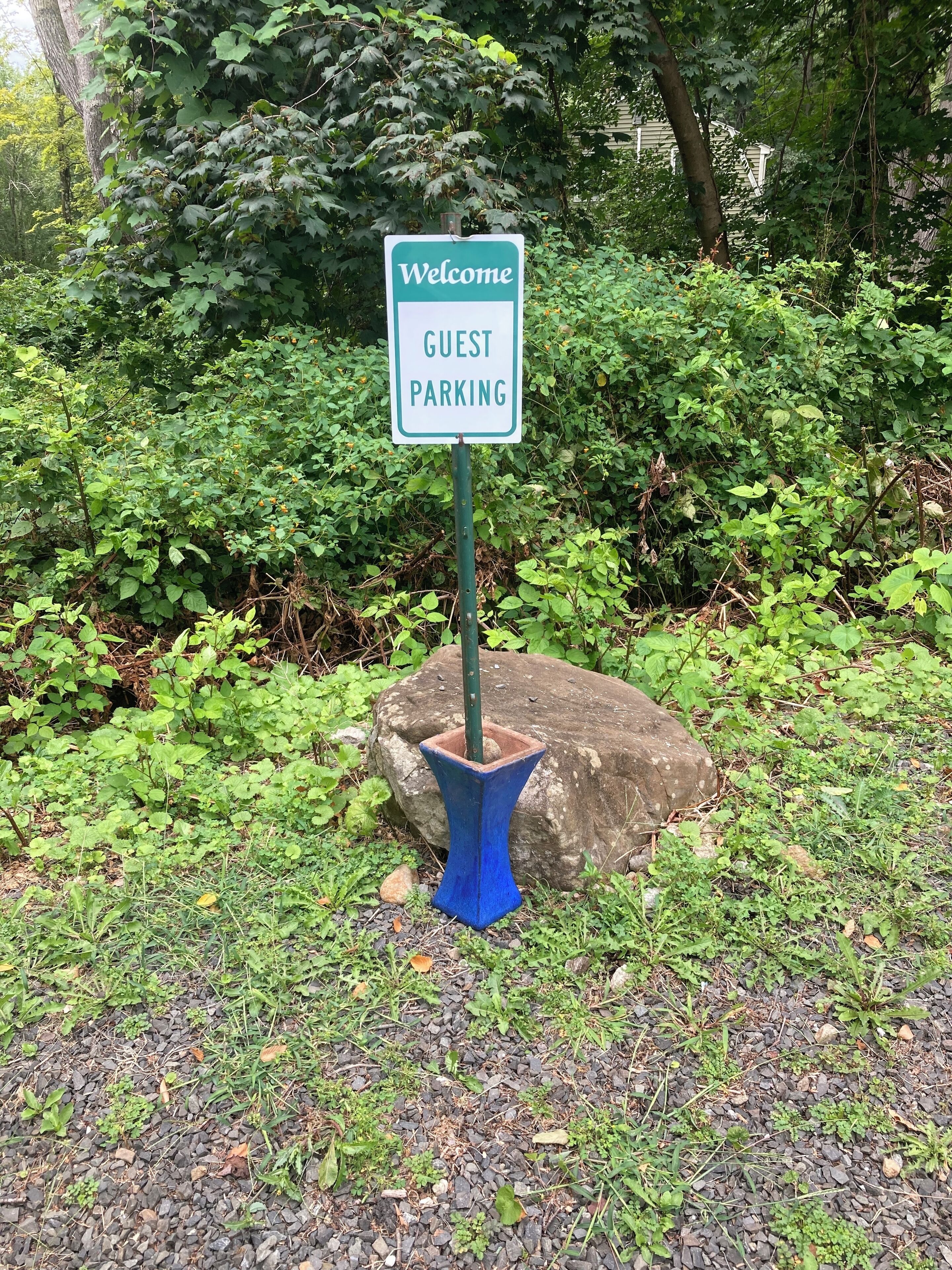 Un panneau indiquant les places de stationnement réservées aux visiteurs, situé dans un espace verdoyant et luxuriant, avec un feuillage dense et un grand rocher à proximité.