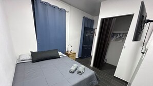 Une chambre avec un lit simple, une couvre-lit grise, un oreiller noir et deux serviettes enroulées. On y trouve une table de chevet en bois avec une lampe, un rideau bleu et une porte qui donne sur une autre pièce.
