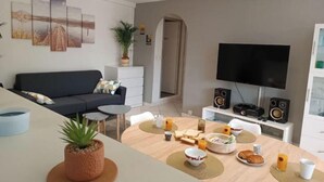 Un salon moderne comprenant un canapé, une table basse, une table à manger avec des aliments pour le petit-déjeuner, un téléviseur et des haut-parleurs.