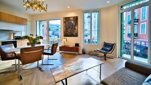 A spacious living room with a modern chandelier, a dining area with a table and chairs, a sofa, and a large window with a city view. - Port of Nice - Scooters Included (Nice)