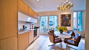 A modern kitchen with wooden cabinets, a dining area with brown leather chairs, and a large window with a view of buildings. - Port of Nice - Scooters Included (Nice)