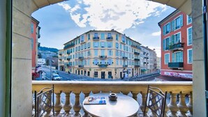 A balcony with a table and chairs, overlooking a multi-story building with balconies and a street view. - Port of Nice - Scooters Included (Nice)