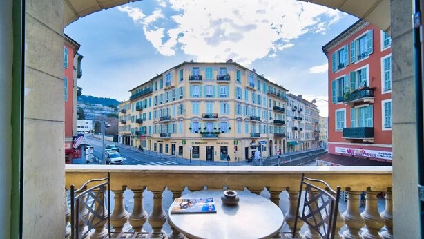 A balcony with a table and chairs, overlooking a multi-story building with balconies and a street view. - Port of Nice - Scooters Included (Nice)