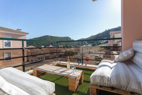 Un balcón con una mesa de madera, una botella y copas, con vistas a una zona residencial con casas y colinas.