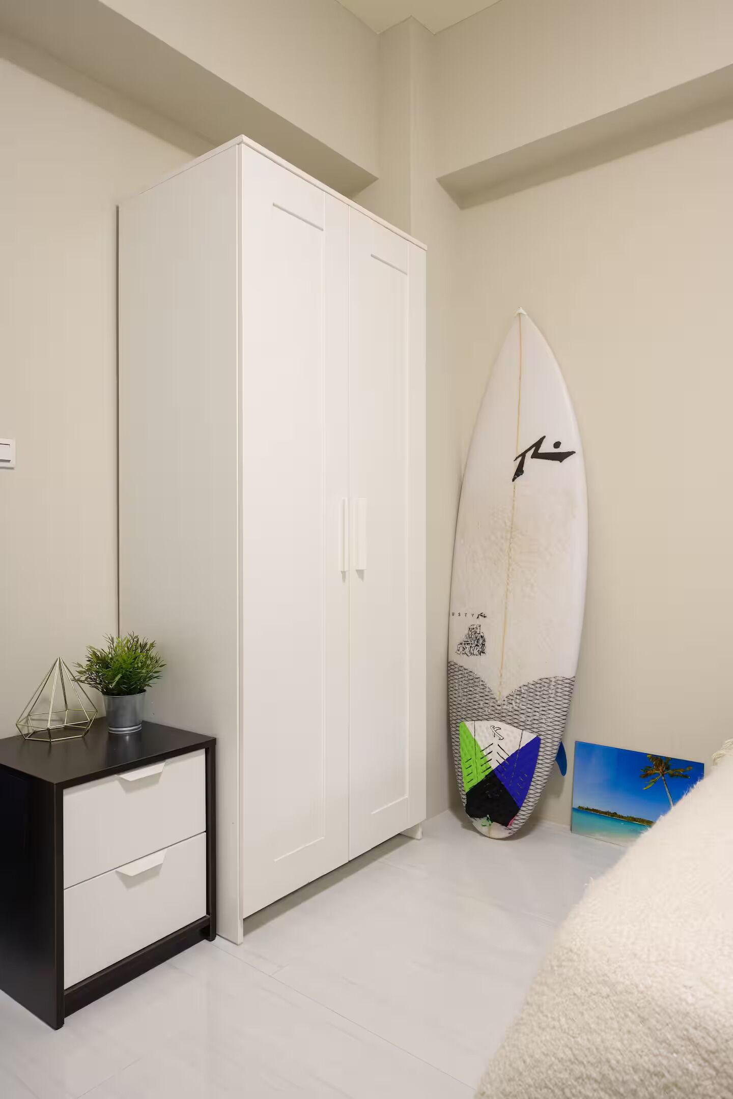 A bedroom with a white wardrobe, a small bedside table with a plant, and a surfboard leaning against the wall.