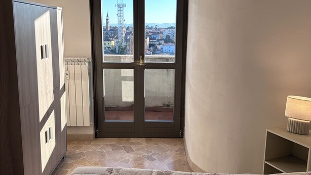 A bedroom with a bed, a wardrobe, a nightstand, and a view of a cityscape through a door. - Mantua Palace (Cremona)