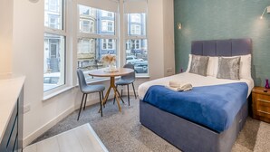 Un dormitorio moderno con una cama grande, un escritorio y una silla. Amplias ventanas ofrecen vistas de edificios.
