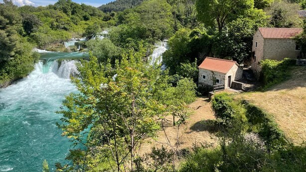 A stone building with a red-tiled roof near a waterfall, surrounded by lush greenery and a clear blue sky. - BUQEZ ECO RESORT - Villa 47 with breathtaking panoramic views directly on the beach. (Pakoštane)