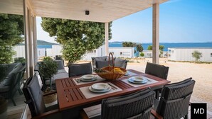A covered outdoor dining area with a table set for two, overlooking a beach and sea. - BUQEZ ECO RESORT - Villa 47 with breathtaking panoramic views directly on the beach. (Pakoštane)