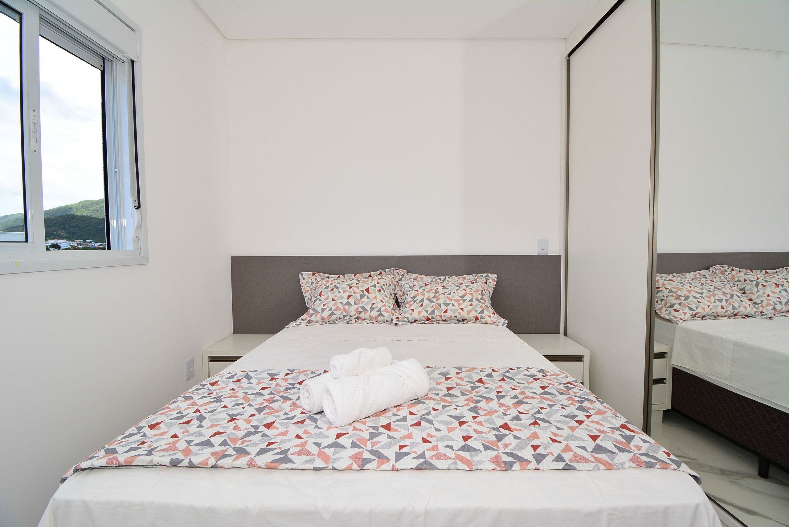 Una cama bien hecha con una colcha estampada y almohadas, una mesita de noche y un espejo grande.