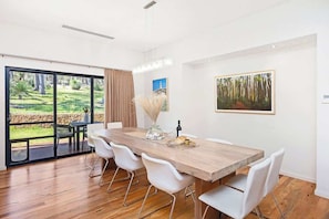 Un coin repas avec une table en bois, des chaises blanches et une vue sur un jardin extérieur, visible à travers des portes vitrées.