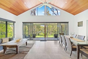 A spacious room with a high ceiling, large windows, a dining area with a glass table, and a view of greenery outside. - Redgum Treehouse - Luxury home in wine country! (Western Australia)