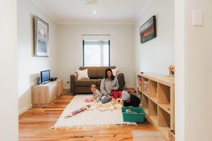Ein Wohnzimmer mit einer Couch, einem Fernseher, einem Kind, das mit Spielzeug spielt, und einem Bücherregal.
