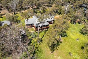 A large, two-story wooden house with a prominent balcony, surrounded by a lush green landscape and trees.