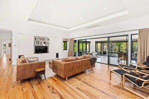 A spacious living area with a wooden floor, a brown sofa, a TV, and a large sliding glass door leading to an outdoor patio. - Redgum Treehouse - Luxury home in wine country! (Western Australia)