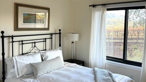 A neatly made bed with white linens and a gray throw blanket, a framed picture on the wall, a bedside lamp, and a window with a view of trees and hills.
