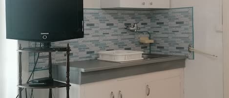 Une petite cuisine avec un mur recouvert de carreaux, un téléviseur posé sur un meuble et une table recouverte d’une nappe à motifs floraux.