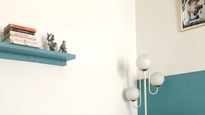 A teal and white armchair with a patterned cover, a small round table with a book, and a shelf with books and figurines. - Charming little art deco apartment (st-claude)