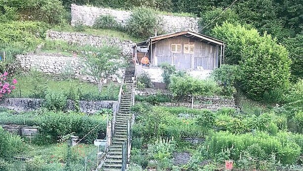 A rustic cabin with a wooden exterior, surrounded by lush greenery and a stone wall. A wooden staircase leads up to the cabin, and there is a small garden area with pink flowers in the foreground. - Charming little art deco apartment (st-claude)
