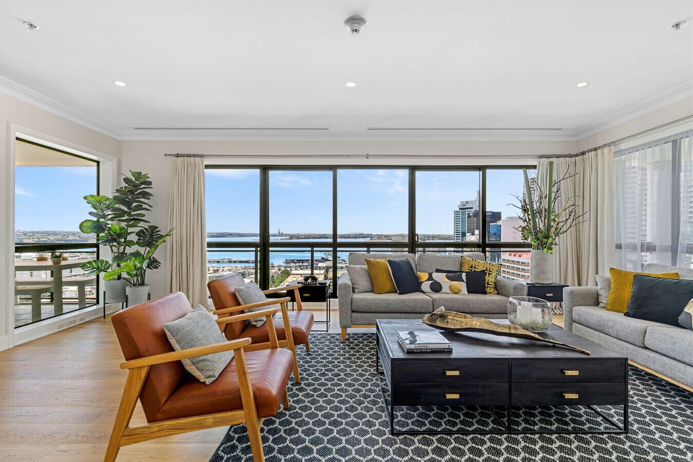 A spacious living room with a large window overlooking a cityscape and water. There are leather armchairs, a sofa, and a coffee table with a magazine.