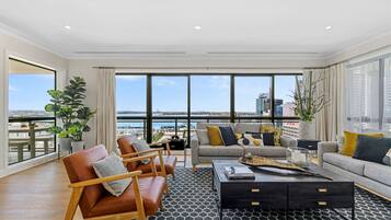 A spacious living room with a large window overlooking a cityscape and water. There are leather armchairs, a sofa, and a coffee table with a magazine.