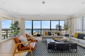 A spacious living room with a large window overlooking a cityscape and water. There are leather armchairs, a sofa, and a coffee table with a magazine.