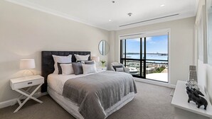 A modern bedroom with a large bed, a nightstand, a desk, and a view of the city through a large window.