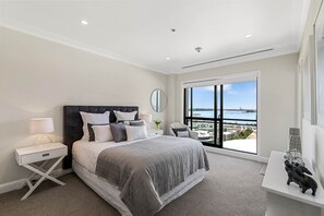A modern bedroom with a large bed, a nightstand, a desk, and a view of the city through a large window.