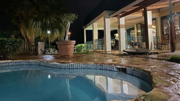 A nighttime view of a swimming pool with a stone edge, surrounded by a covered patio area with a railing. - Casona Mirimou-Country and Beach House! (Luquillo)