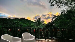 A wooden deck with white plastic chairs and a railing, overlooking a lush green landscape with a sunset in the background. - Casona Mirimou-Country and Beach House! (Luquillo)