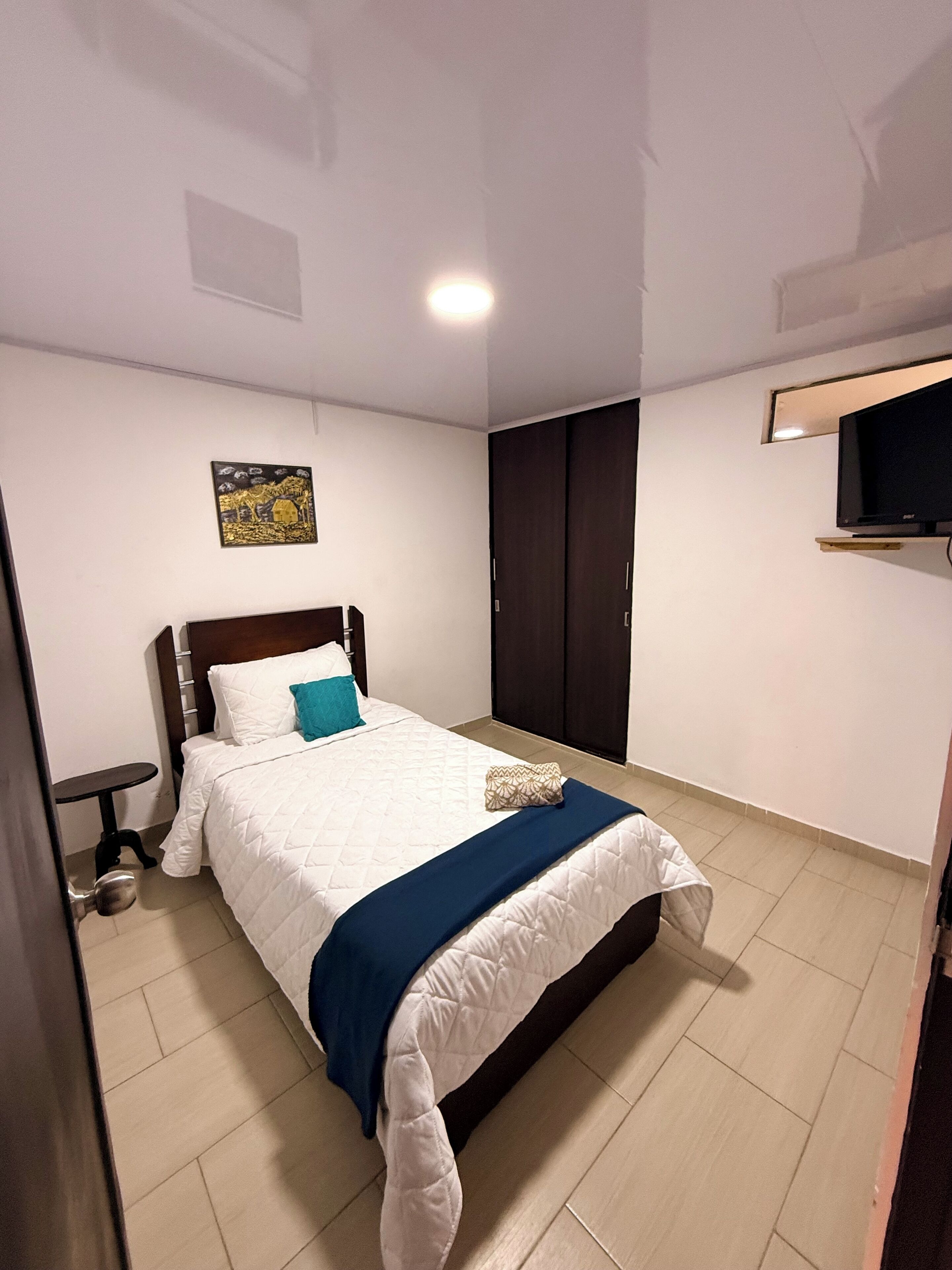 Una cama bien hecha con una manta azul, una mesita de noche pequeña, un televisor montado en la pared y una puerta con pomo.