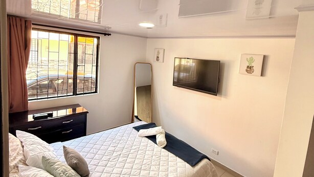 Ein Schlafzimmer mit einem Bett, einer Kommode, einem an der Wand befestigten Fernseher und einem Fenster mit Vorhängen.
