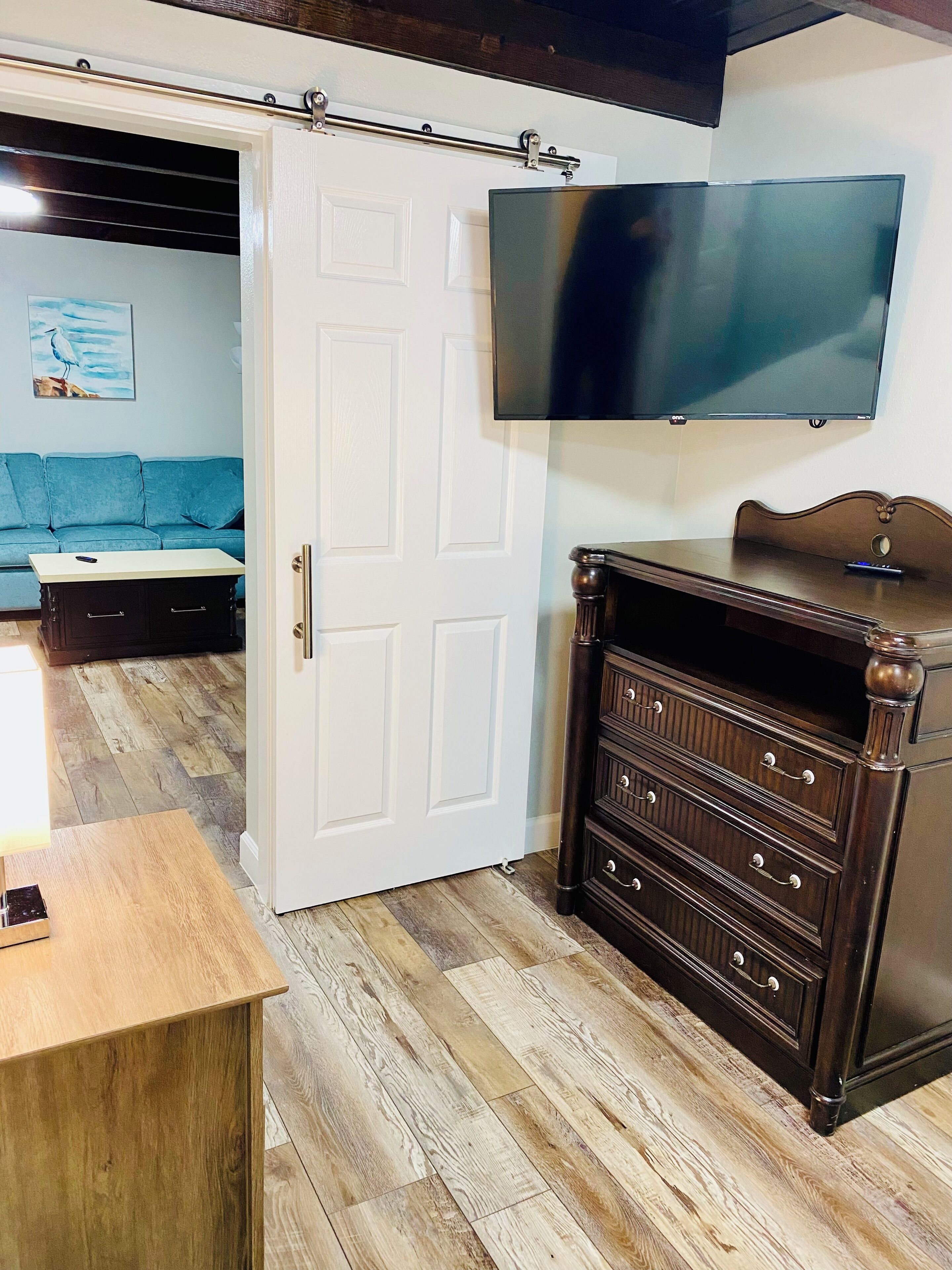 A room with a wooden floor, a white door, a dark wooden dresser, a blue sofa, and a flat-screen TV mounted on the wall.