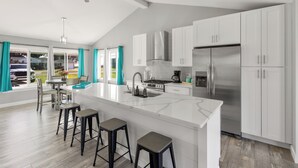 A modern kitchen with white cabinets, a large island with stools, a stainless steel refrigerator, and a ceiling fan. - Charming 3-bedroom house on the Canal! Fireplace! Firepit! Pets Welcome! (Harrison Twsp)