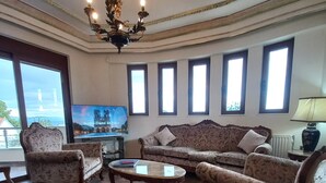 A living room with a sofa, armchairs, a coffee table, and a chandelier. There are windows with blinds and a television on the wall. - Salty Sea Breeze nearby Athens airport (Artemis)