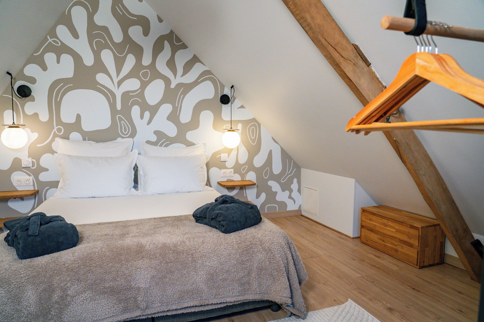 A bedroom with a sloped ceiling, a bed with white linens, two bedside lamps, a wooden dresser, and a hanging rack.