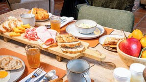 A breakfast setting with a variety of foods, including bread, pastries, fruits, and beverages, arranged on a wooden table in a cozy room with a fireplace.