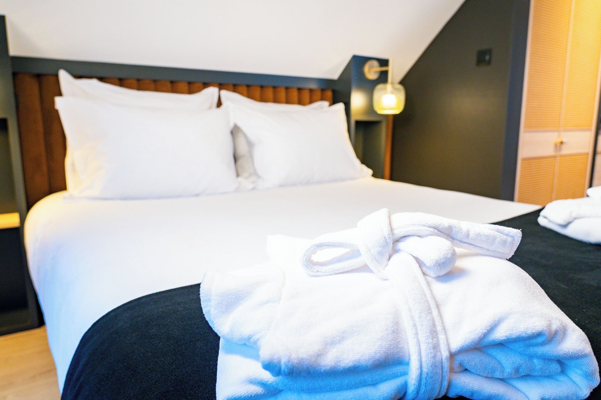 Una cama bien hecha con sábanas y almohadas blancas, cabecera de madera y una lámpara fijada a la pared.