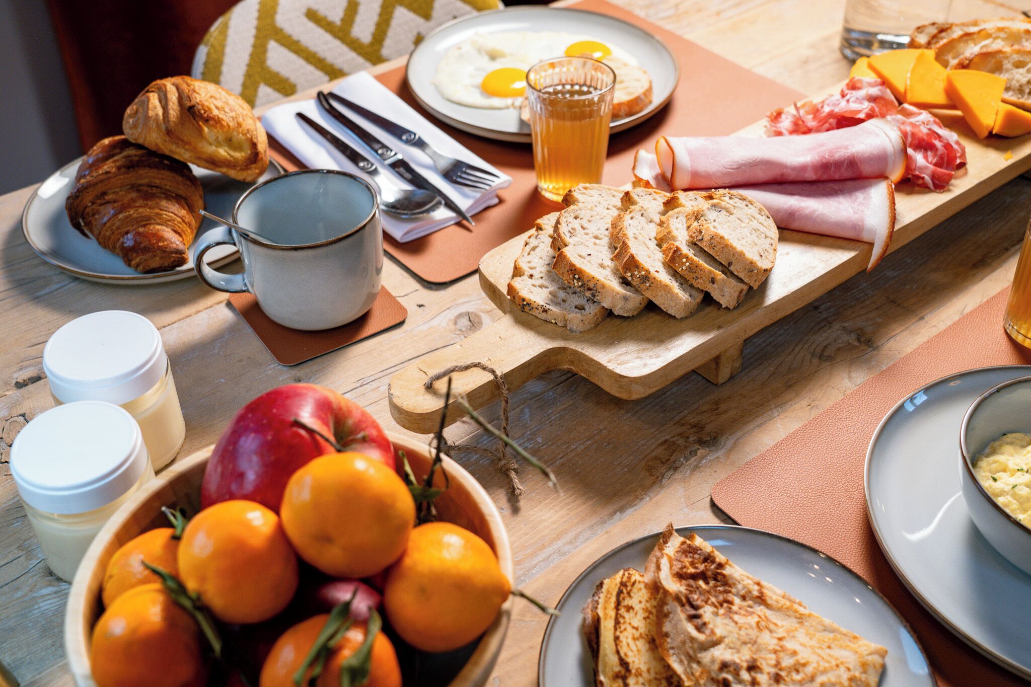 Une table de petit-déjeuner avec des croissants, des œufs, du fromage, du pain et un bol d’oranges.