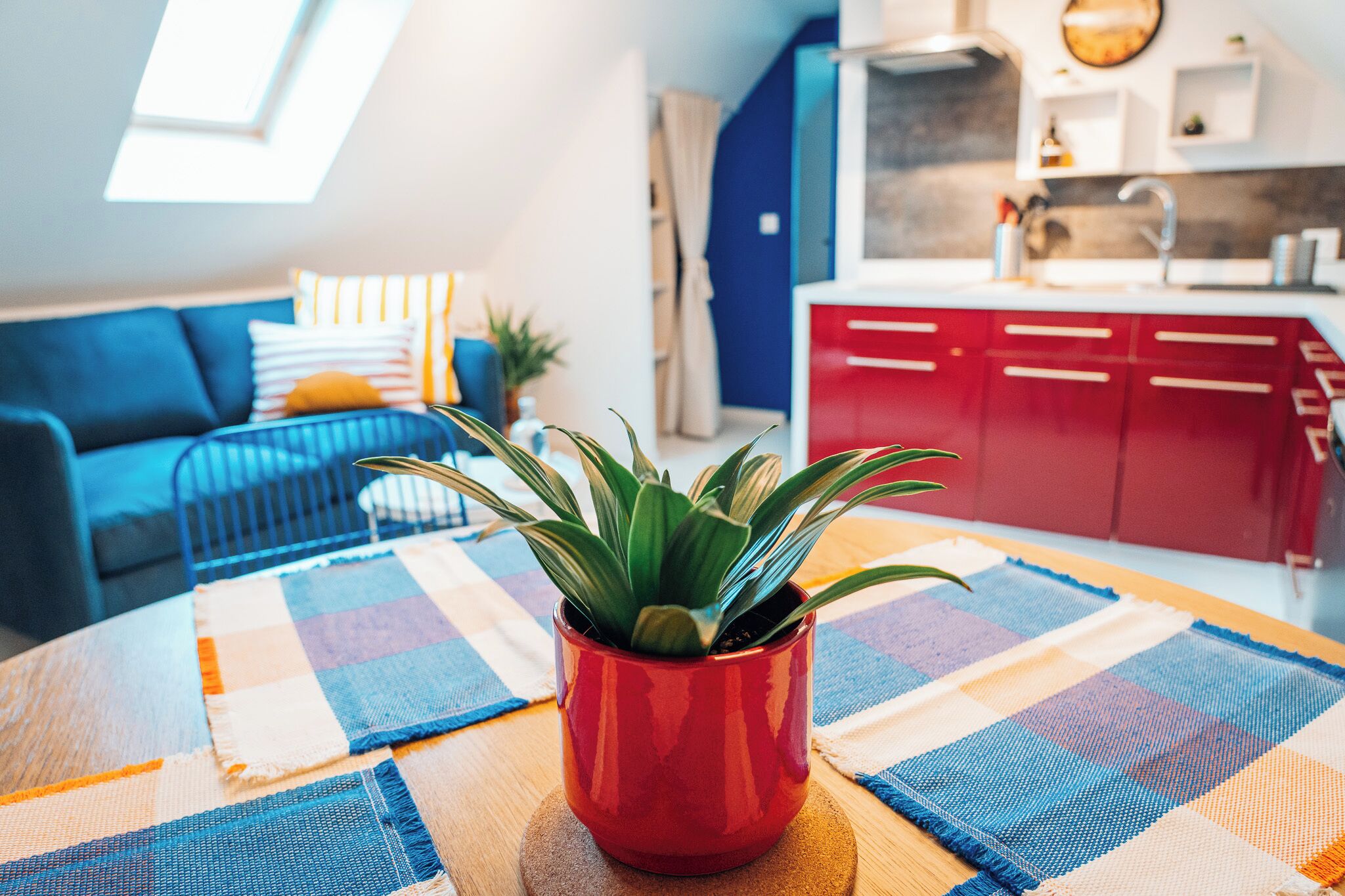 Un salon chaleureux avec un canapé bleu, un meuble rouge et une plante en pot posée sur une table ronde recouverte d’un chemin de table à carreaux.