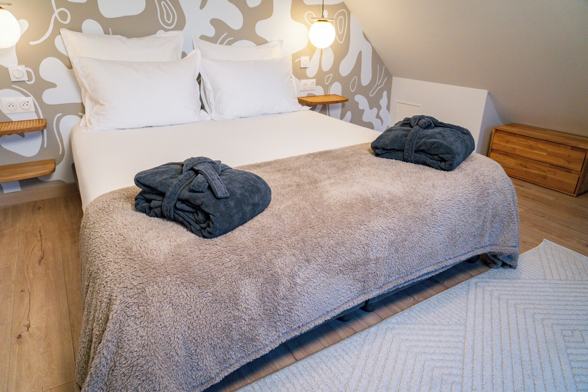 A neatly made bed with a fluffy blanket, two folded robes on the bed, a bedside table with a lamp, and a wooden nightstand with a power outlet.